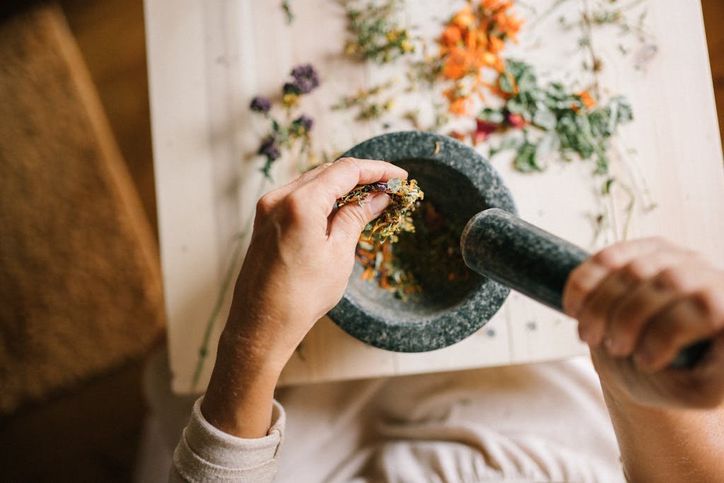 Top view of hands grinding dried herbs using a mortar and pestle on a wooden table.